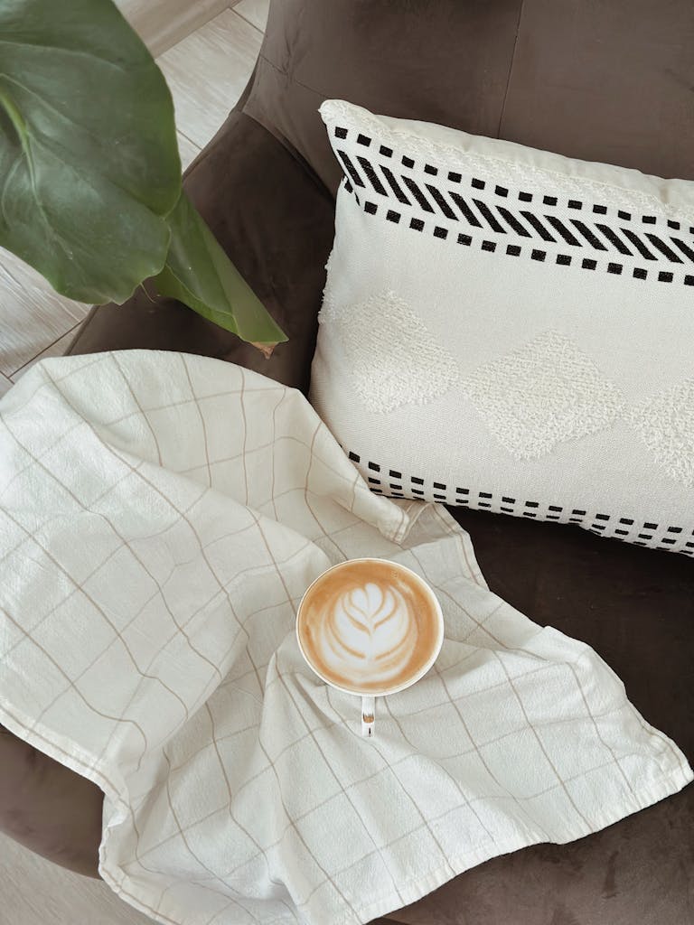A warm cappuccino with latte art on checkered cloth and comfy pillow in cozy setting.