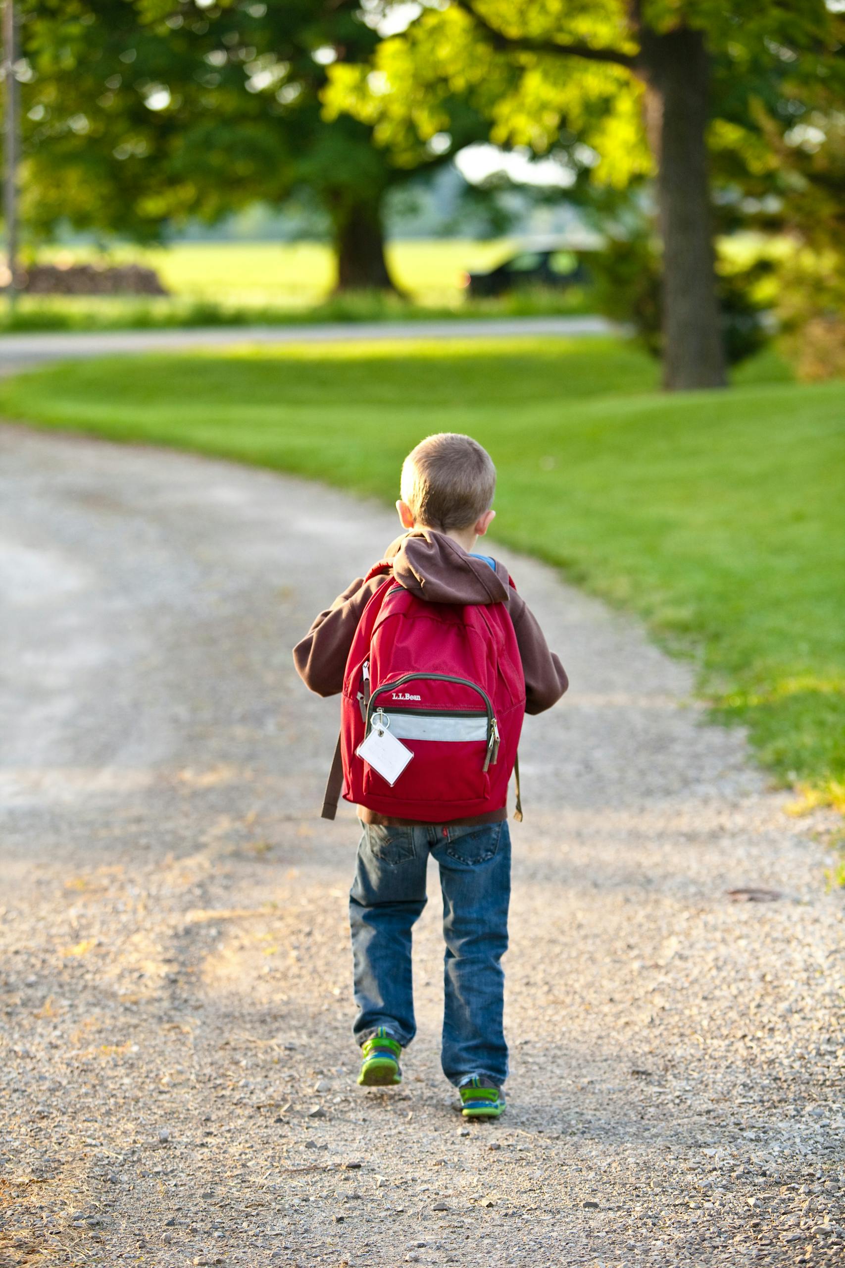 A young boy with a red backpack walking on a gravel path in a sunny park setting.