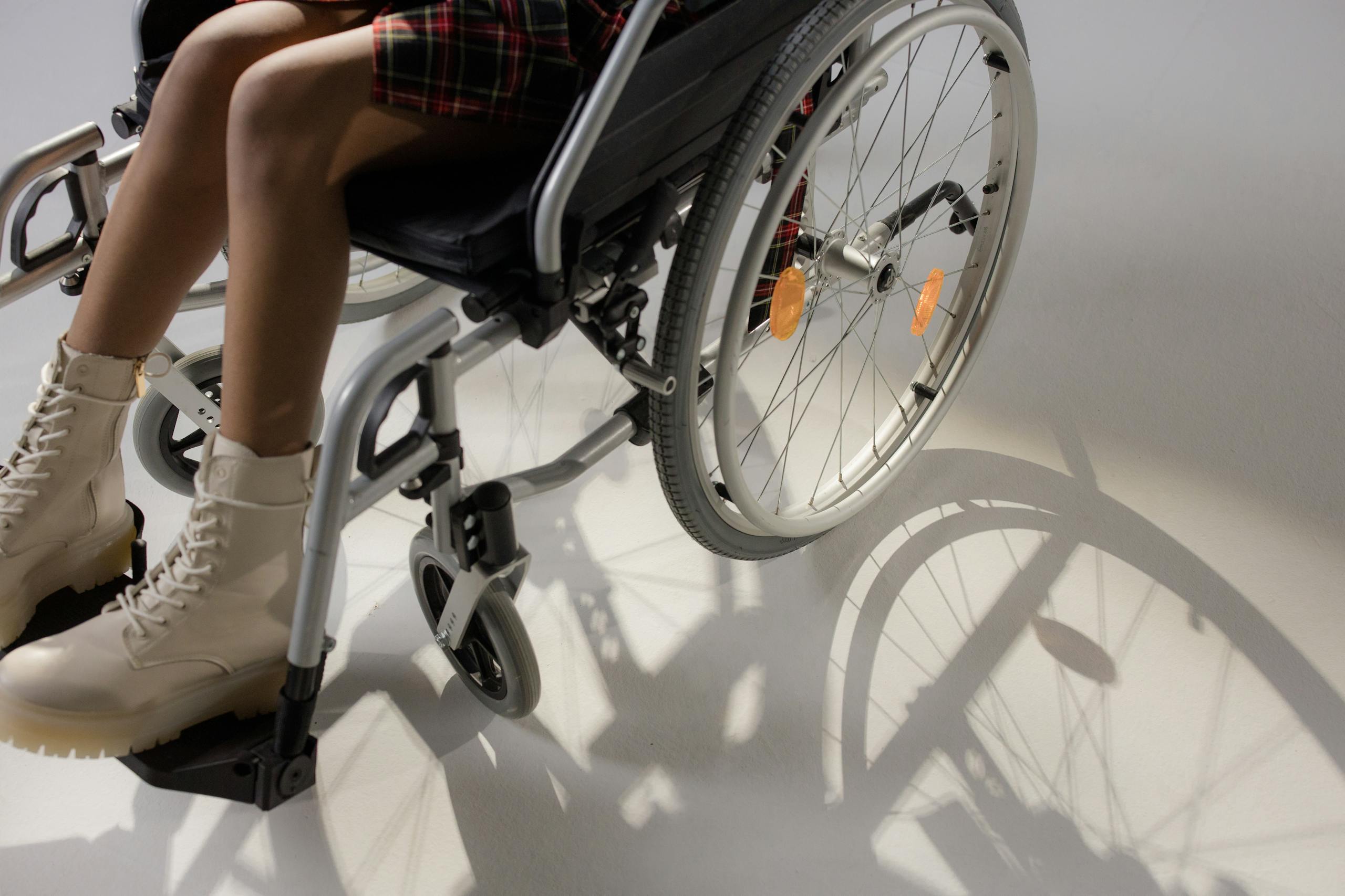 A young woman sitting in a wheelchair, wearing stylish beige boots, casting a shadow.